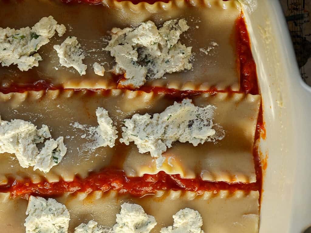 Top view of uncooked lasagna in a baking dish, featuring wavy pasta sheets partially covered in red tomato sauce and dollops of white ricotta cheese mixture.